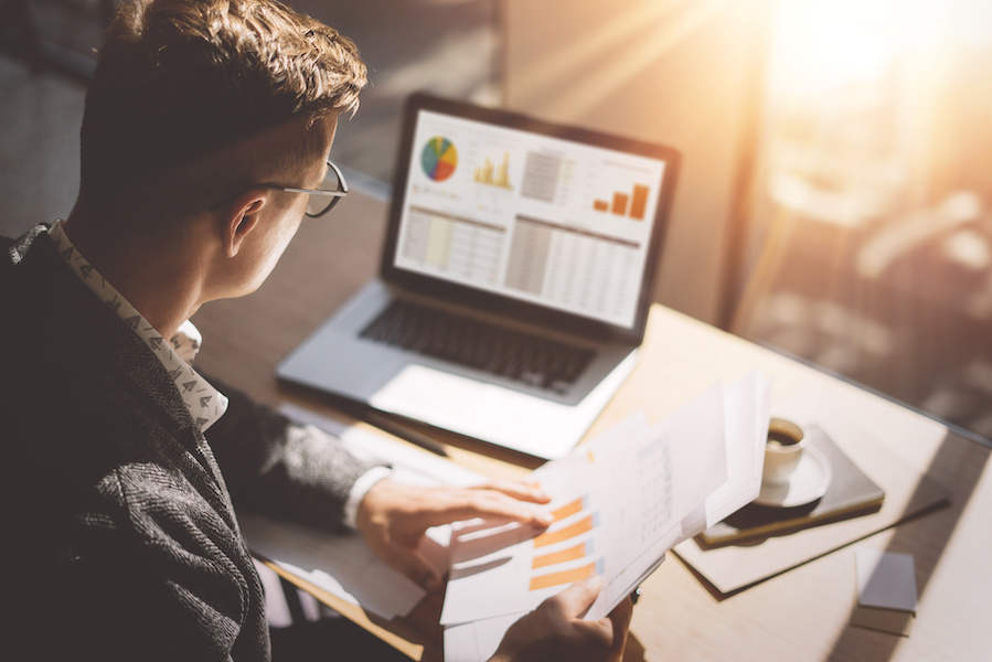 Young finance market analyst in eyeglasses working at sunny office on laptop while sitting at wooden table.Businessman analyze document in his hands.Graphs and diagramm on notebook screen.Blurred